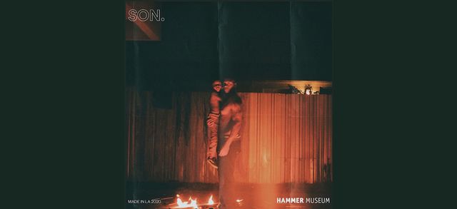 Dark photograph outside, at nighttime, of a man holding a small child, while standing in front of a fire in the foreground. The man is looking at the viewer, and the fire reflects against a fence in the background. Words are on the image: "SON." in the top left corner, "Made in L.A. 2020" in the lower left corner, and "Hammer Museum" in the lower right corner.