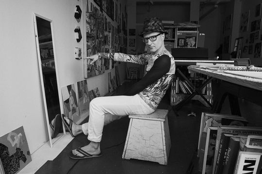 Black and white photo of artist Wayne Koestenbaum seated and pointing at himself in a mirror while looking at the camera