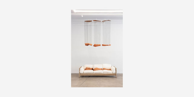 Broken terra cotta forms lay on a white sofa, as three other forms hang from the ceiling above