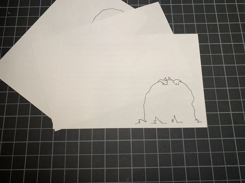 Pencil drawing of two paws on top of a rock with ears peeking out on a white card. Black and white grid in the background.