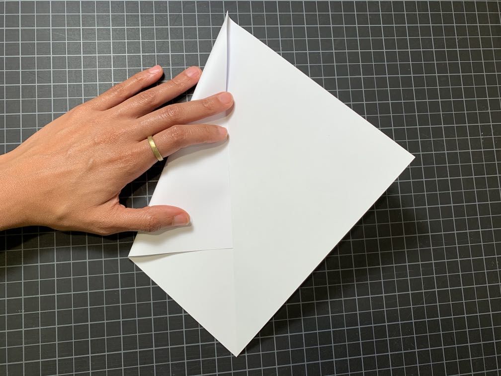 A diamond shaped piece of paper with a crease down the middle being folded with the left point coming into the center crease