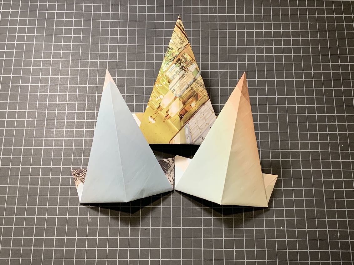 Three folded triangle shaped papers, one blue, one multiple shades of yellow, one light pink and white