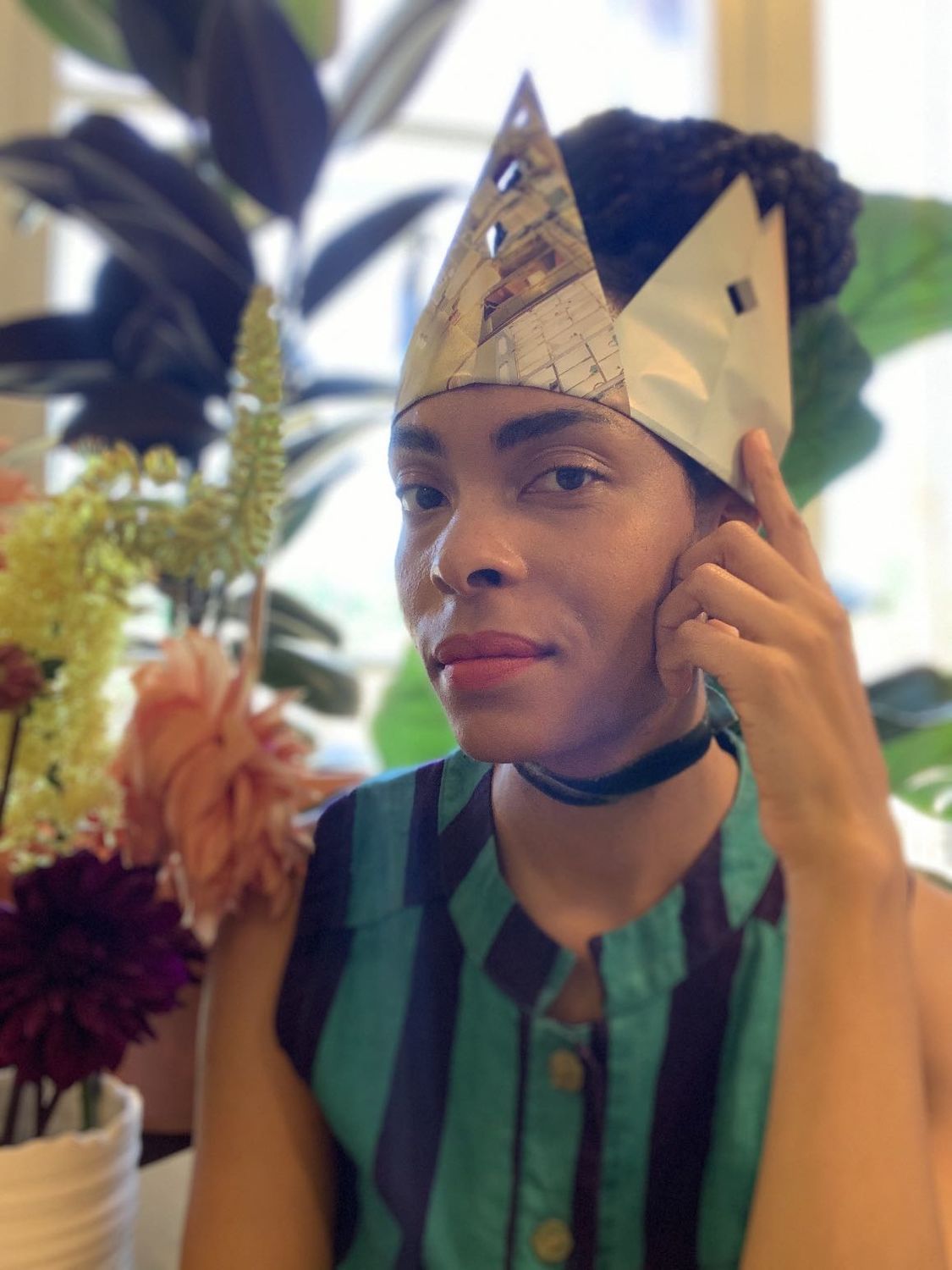 Kenturah Davis wearing a green and purple striped sleeveless top and mult-colored handmade paper crown, with multiple plants in the background.