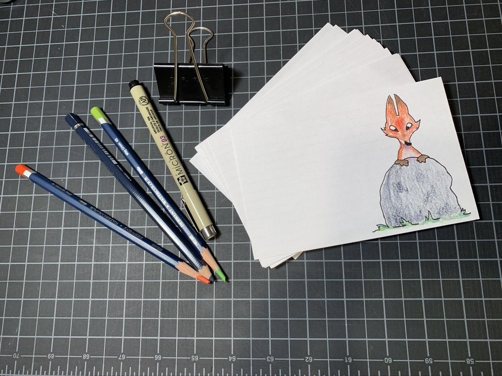 Three colored pencils, one marker, and one file clip to the left of a stack of white cards. The top card has a drawing of an orange fox peeking from behind a gray rock.