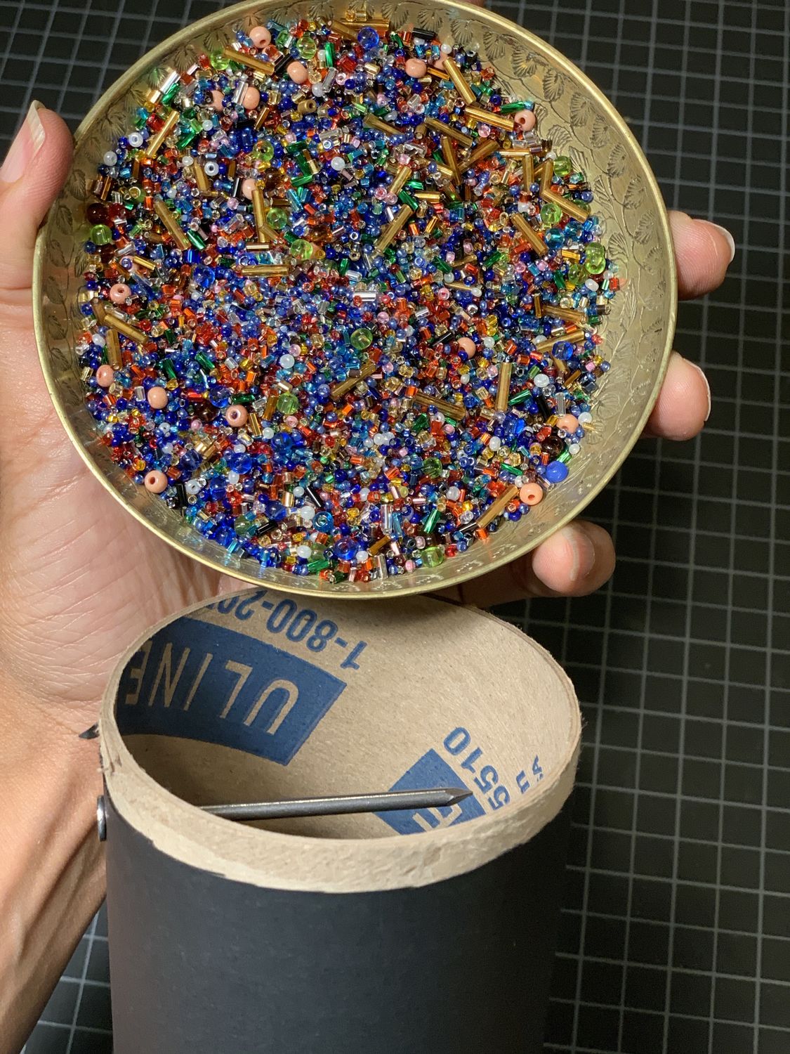 Multi-colored beads in a small gold dish held above the opening of a tube covered in black, one silver nail visible at the tube's opening