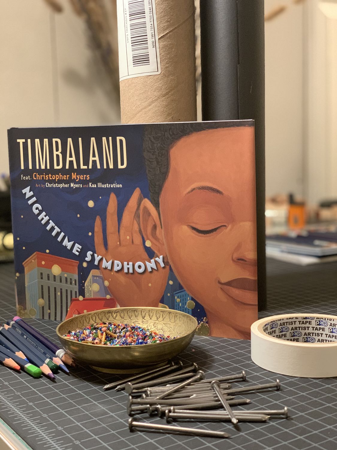 Cover of the book "Nighttime Symphony" with a gold bowl of multi-colored beads in front, multi-colored colored pencils on the left, silver nails in the foreground, cardboard tubes behind the book, and masking tape to the right.