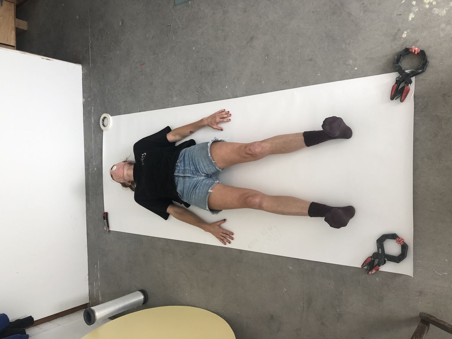 Woman in black shirt, blue jean shorts, and black socks laying on her back on top of a white piece of kraft paper