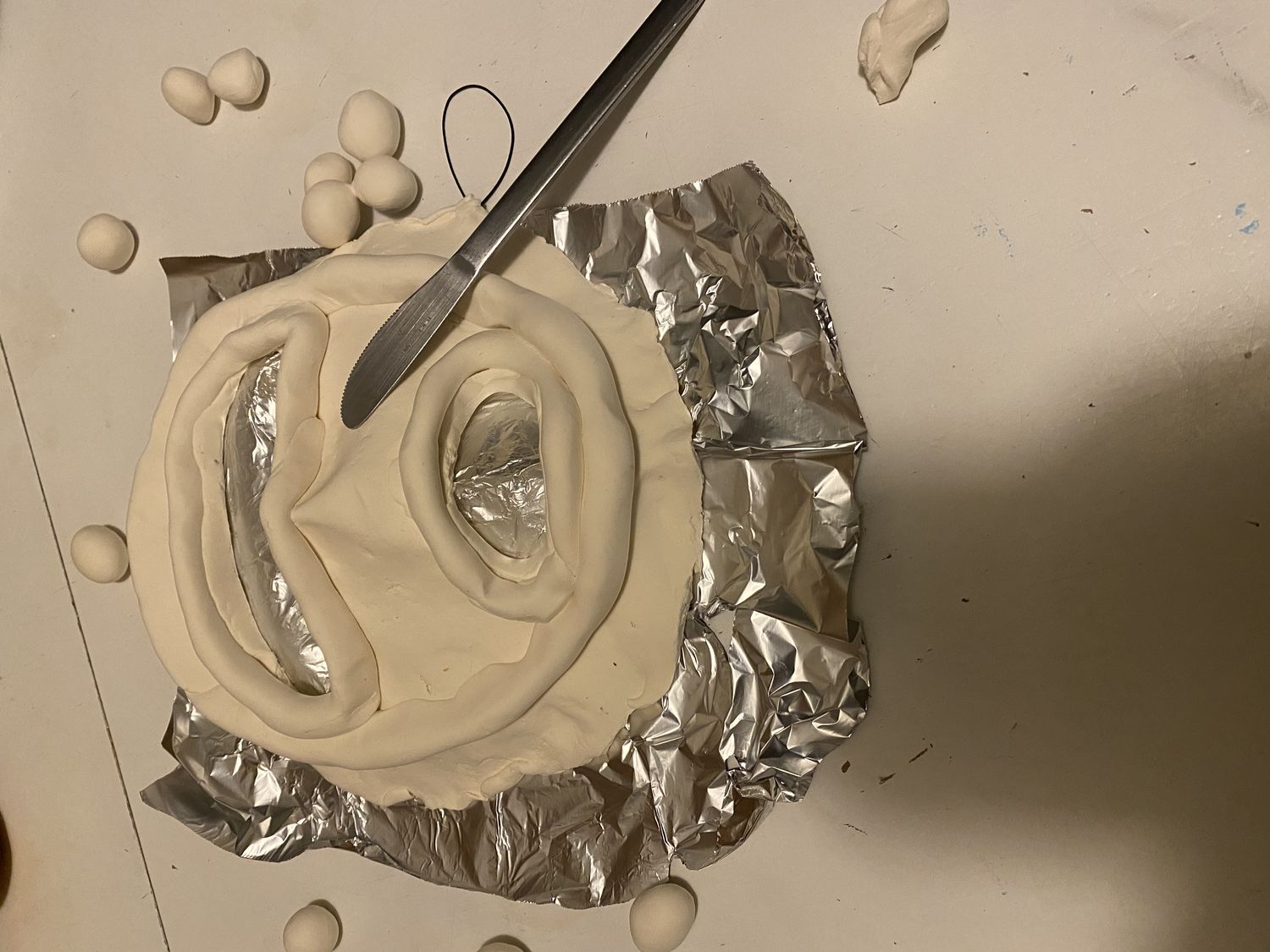 Clay mask with mouth and eye area hollowed out laid on top of piece of foil on flat surface. Kitchen knife laid across mask with small balls of clay around the object