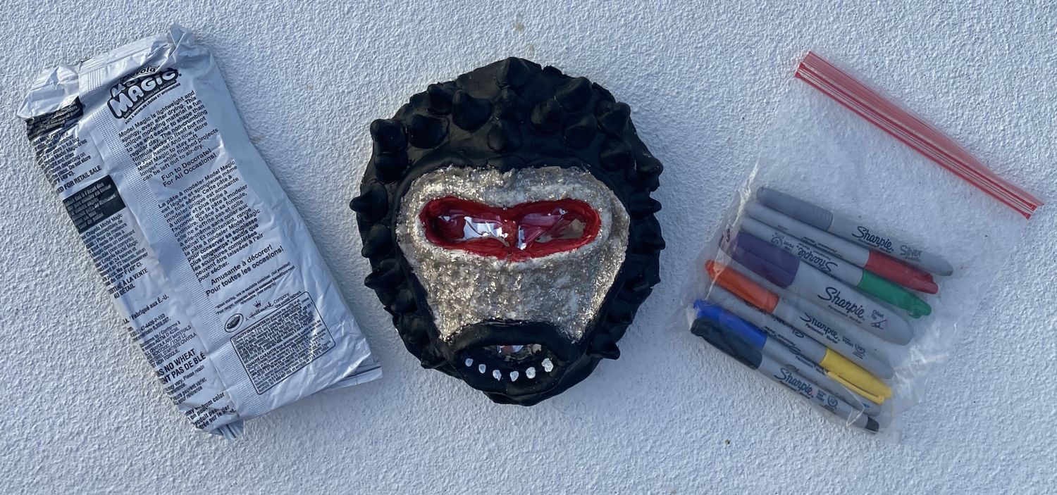 One pack of Model Magic, one clear plastic bag with markers, and a mask with red eyes, gold face, and black hair all around pictured in the center