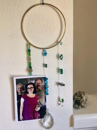 Hanging suncatcher, large gold hoop with three strings of beads hanging at the bottom. String on the left has a photograph featuring three smiling people