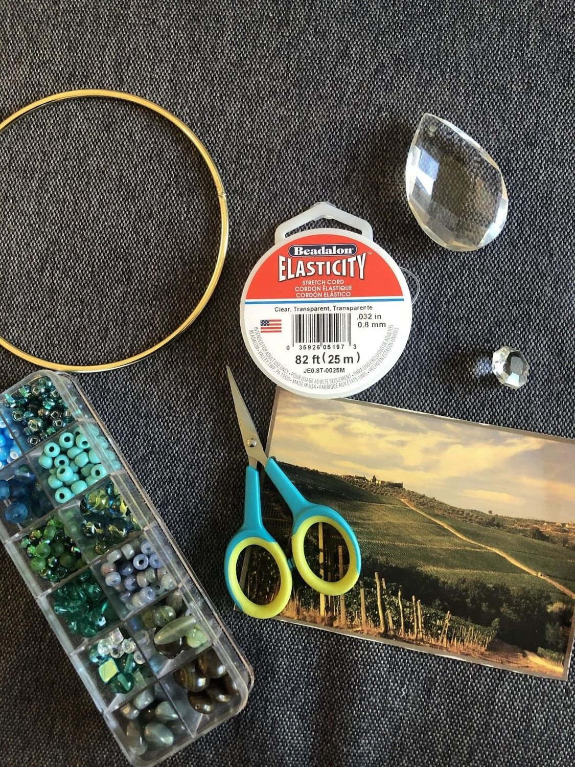 Supplies for creating a suncatcher laid across a flat surface, including gold hoop, plastic container of small blue and green  beads, kids scissors, a photograph of a landscape, and a large plastic gem.