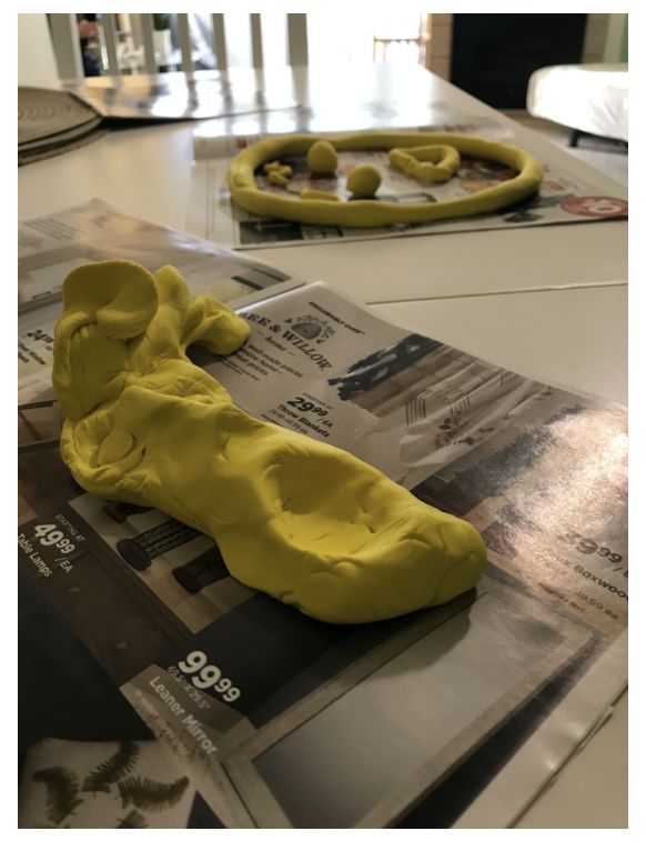 Two yellow sculptures. In the foreground a face with an open mouth and tall, curvy hair. In the background a smiley face. Both laid on top of a flat surface.