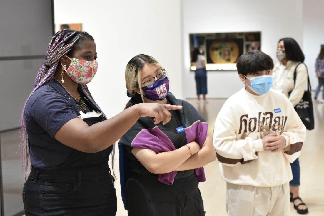 alea, who has brown skin, purple braids, and is wearing a multi-colored mask, points out something in the galleries to two people standing to her left