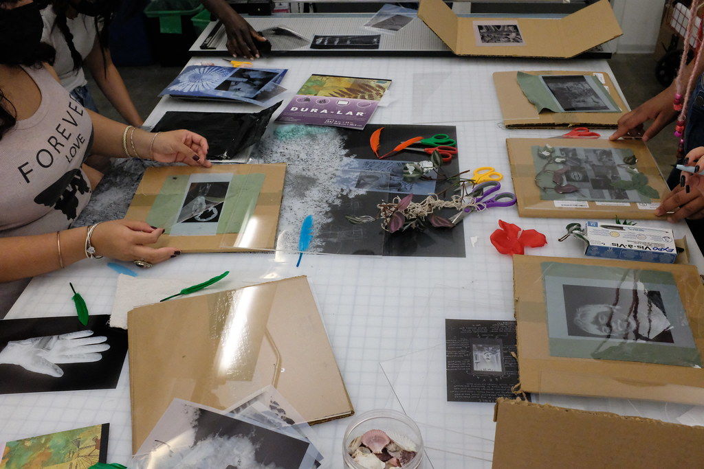 Overhead image of a table covered in artmaking supplies, with the hands of a few students making cyanotypes