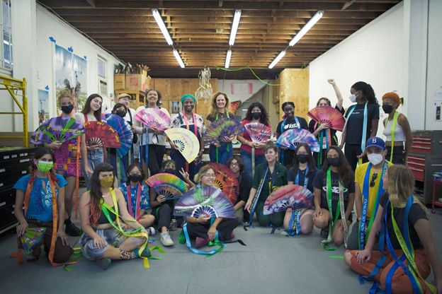 A group of students surround the artist Andrea Bowers, holding colorful paper fans and wearing ribbons