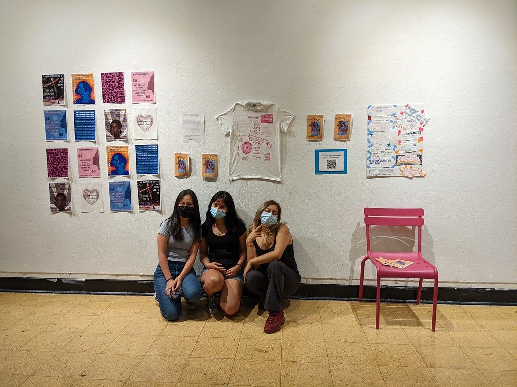 Photograph of three young women kneeling in front of a wall of artworks by Andrea Bowers