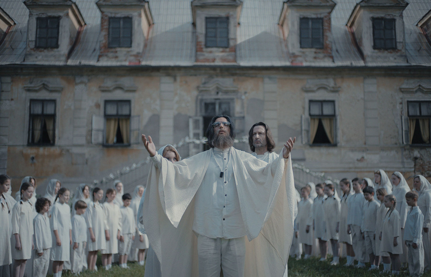 A man dressed in white holds his hands in the air, with a large group of children dressed all in white behind him