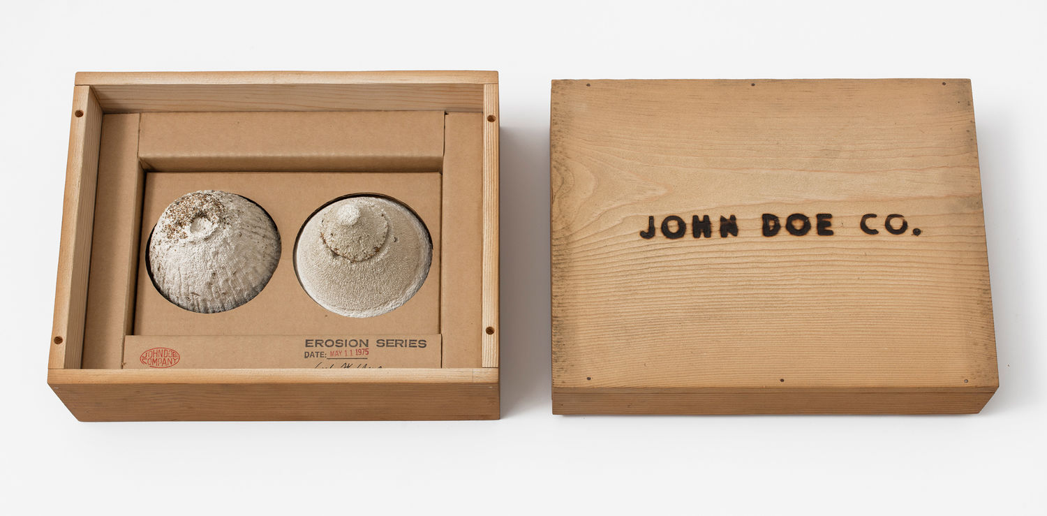 A simple wooden box: on the left, two small mounds of sand sit in the box, with a stamp reading "EROSION SERIES," on the right, the box top with the words "JOHN DOE CO." burned into it