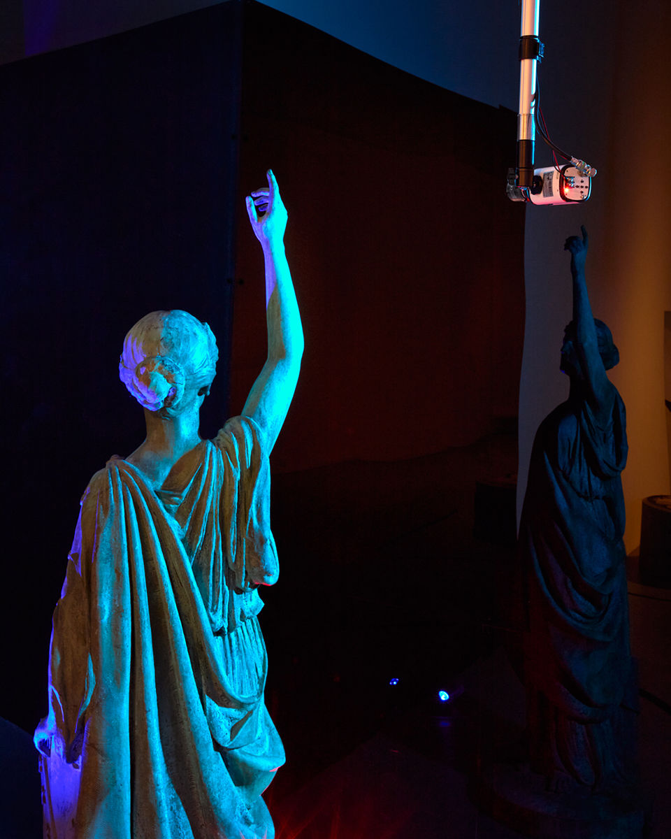 A bronze sculpture of a female figure lifting her right arm, relfected in a black wall, with a surveillance camera overhead