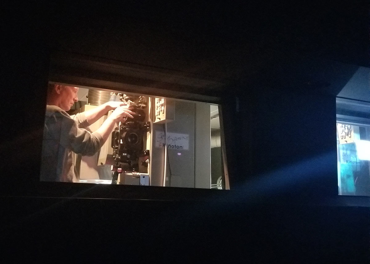 The projectionist loads a reel onto the projector on the left, while the previous reel projects onto the screen through the window on the right. 