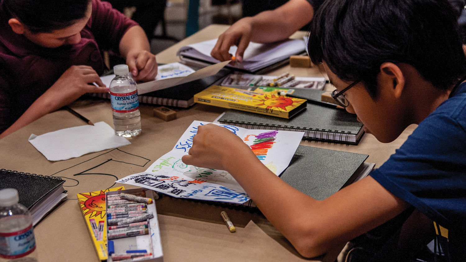 Two children sit across a table and draw on paper