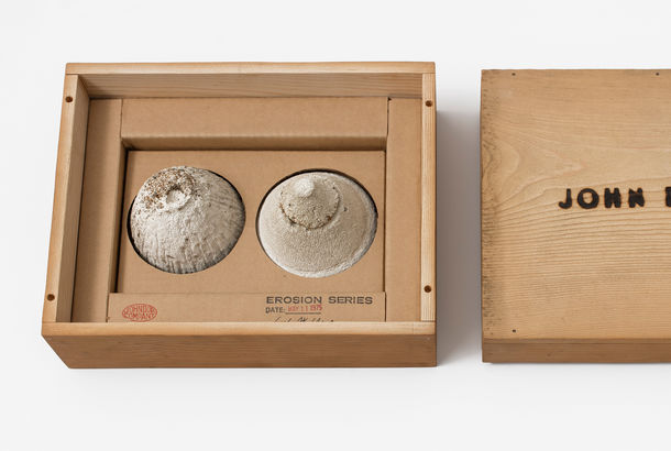 A simple wooden box: on the left, two small mounds of sand sit in the box, with a stamp reading "EROSION SERIES," on the right, the box top with the words "JOHN DOE CO." burned into it