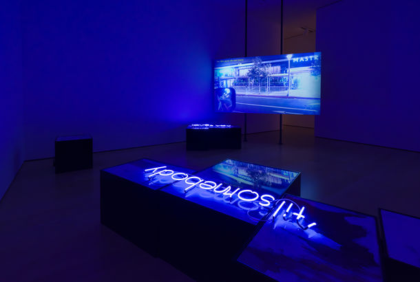 Installation with a video screen in the background showing a street scene, and a neon sign reading "til' somebody" laying on a plinth in the foreground