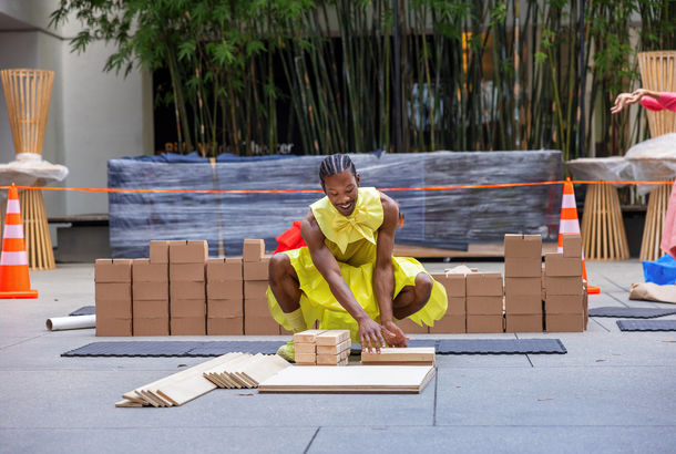 A dancer in a yellow outfit smiles as he kneels in front a stack of wood pieces
