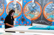 Visitor in a gallery stands, looking down at a device embedded in a table. A very large teal book is also on the table. In the background is a wall-sized painting with geometric designs, red and blue background with black lines.circles, 