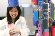 A young girl stands, smiling, wearing a hat made of blue construction paper, a white coat, and silver glitter on her fingers. To the right of her is a structure made of rolls of colored construction paper with words and tinsel.