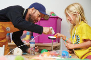 A girl in a yellow shirt is holding up an art project and is assisted by a man in a baseball cap who leans over with a glue gun pointed at the girl's project.