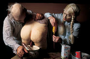 Two people wearing doll-like masks lean over a table where a nude bottom that appears made from plaster or papier-mache is sitting,  Various bowls and pots sit on the table as well.  