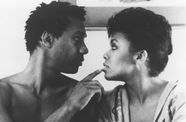 Still from the film The Brother From Another Planet (1984) showing a close-up of a shirtless man looking into the eyes of a woman in a robe and touching her lip