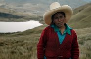 A woman stands outside against a hilly, green landscape with a lake  or river in the background. The woman wears a large, white cowboy hat and wears a red cardigan over a blue shirt.