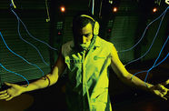 Still from the film Sleep Dealer (2008) showing a man in a face mask with his arms spread wide and connected to glowing tubes