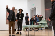 Two young man stand in a gallery next to a sculpture among a crowd