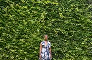 A woman in a dress stands facing the camera  in front of a wall of plants