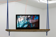 An open laptop computer sits on a wooden shelf that hangs from the ceiling. The walls in the background are white, and the edge of an open skylight are visible at the top right of the image.  