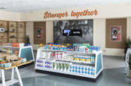 Photograph showing inside of a grocery store, with low shelving. On the back wall the words "Stronger Together" are written in orange cursive. Below this a black and white photographic mural, with two video monitors mounted over top.
