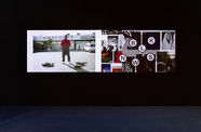 A photograph of two videos projected side-by-side against a black wall that takes up two-thirds of the image. The carpet in the lower third of the photograph is blue.