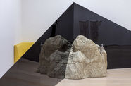 A view of the corner of a gallery. Two dark triangular structures extend across the corner, meeting in the middle at the tall ends. A large rock sits on the floor in front of the triangles, where they meet. A drinking fountain spout extends from the  top of the rock. Behind the triangles, between them and the wall, the edge of a yellow piece of furniture is visible.