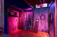 The corner of a room in a haunted house, with whips hanging on a wall lit in purple lighting
