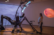 A person stands in a dimly lit gallery with abstract video projections on the walls, wearing headphones connected to a mechanical sculptural installation by the artist Sondra Perry 
