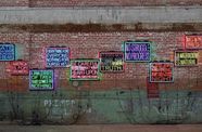 Photo of neon light artworks installed on a brick wall. The neon artworks are signs with phrases in Spanish and English related to immigration.