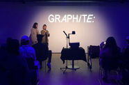Photo of a dark room with the farr wall spotlit and the word "GRAPHITE:" projected on the wal in white letters. On the left and right we see the backs of people sitting in chairs. Two women stand against the far wall and speak into a microphone. 