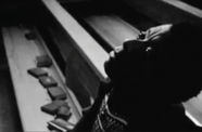 Black and white photograph, with a person in the foreground, visible from the chest up, sitting in a church pew and looking upwards. The person has dark skin and short dark hair. The pew extends diagonally into background at the upper left corner. Books are strewn on the pew next to the person. 