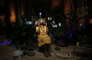 A person sits in a beauty salon chair in a dark room with potted palm trees behind her. The person has medium-dark skin, wears a yellow dress with several metal necklaces, is barefoot, and holds the chair's drying helmet over their face with two hands. Various-sized video monitors peek out rom the plants. 