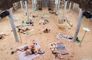 Photograph of people in bathing suits laying on a sandy ground inside of a building with columns. 