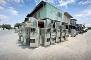 A neat wall of silver air conditioners are stacked three-high on a dusty street with a teal building in the background