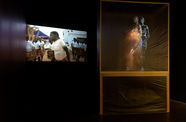 A square structure covered in clear film obscures a figure inside, while a projection plays on the wall to the right